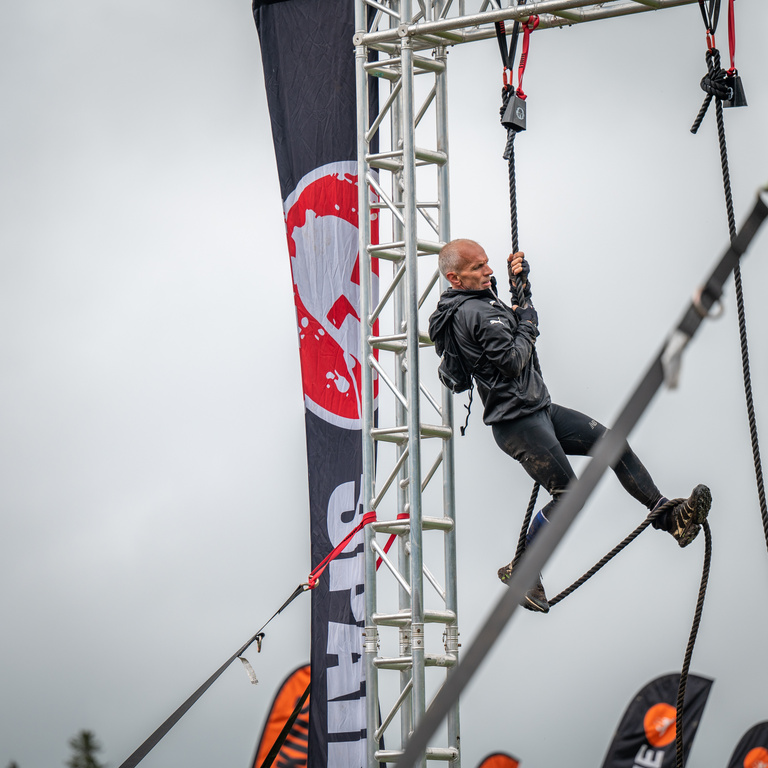 An athlete climbs a hanging rope beside a tall metal obstacle structure.
