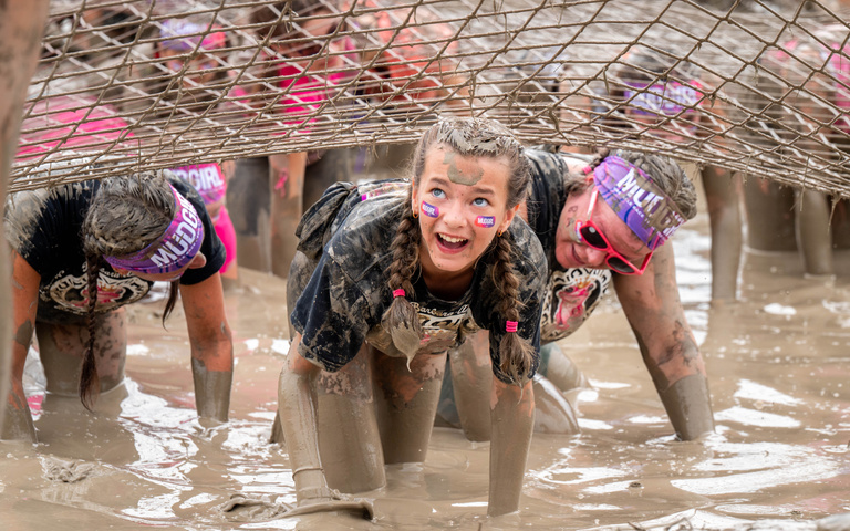 A smiling participant makes their way through a muddy obstacle.