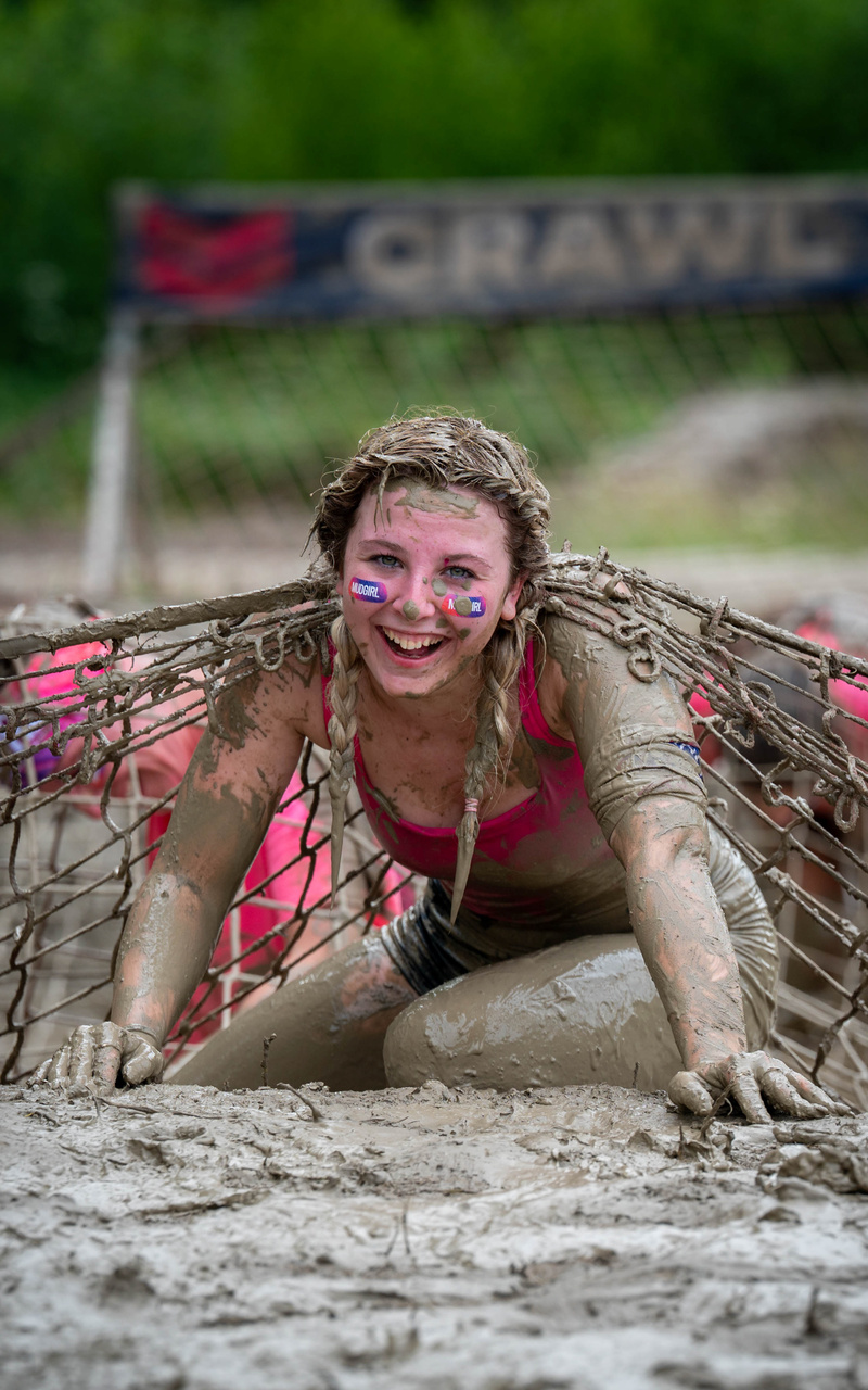 A smiling participant crawls toward the camera through mud beneath a rope net.