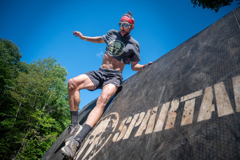 An obstacle racer climbs a curved wall beneath a bright blue sky.