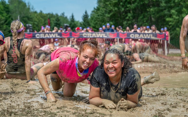 Two smiling participants crawl side by side through a muddy obstacle.