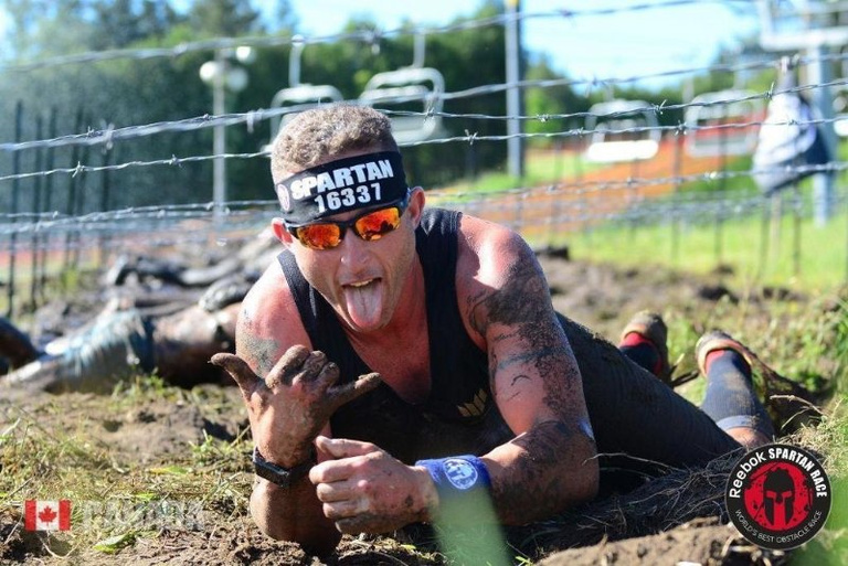 A racer wearing sunglasses crawls through mud beneath a wire obstacle.