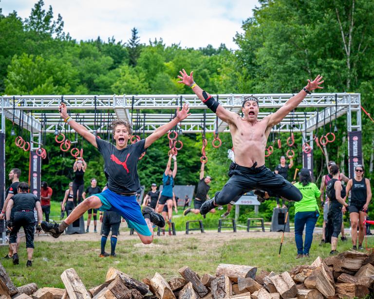 Two athletes jump with their arms raised in front of an outdoor obstacle course.