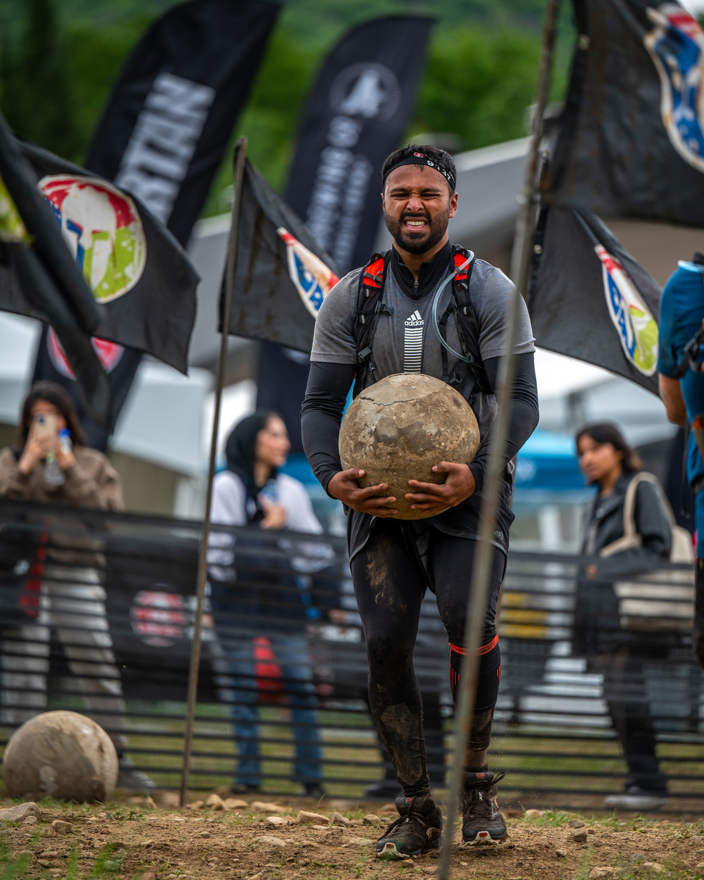 An athlete carries a heavy round stone across an outdoor obstacle course.