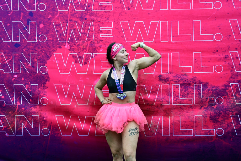 A participant wearing a medal and pink skirt celebrates in front of an event backdrop.