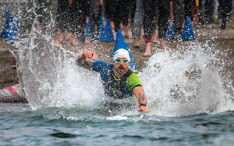 A swimmer charges into the water amid a wall of spray and other competitors.