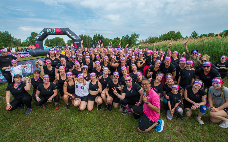 A large group of smiling participants poses together outdoors in front of an event finish arch.